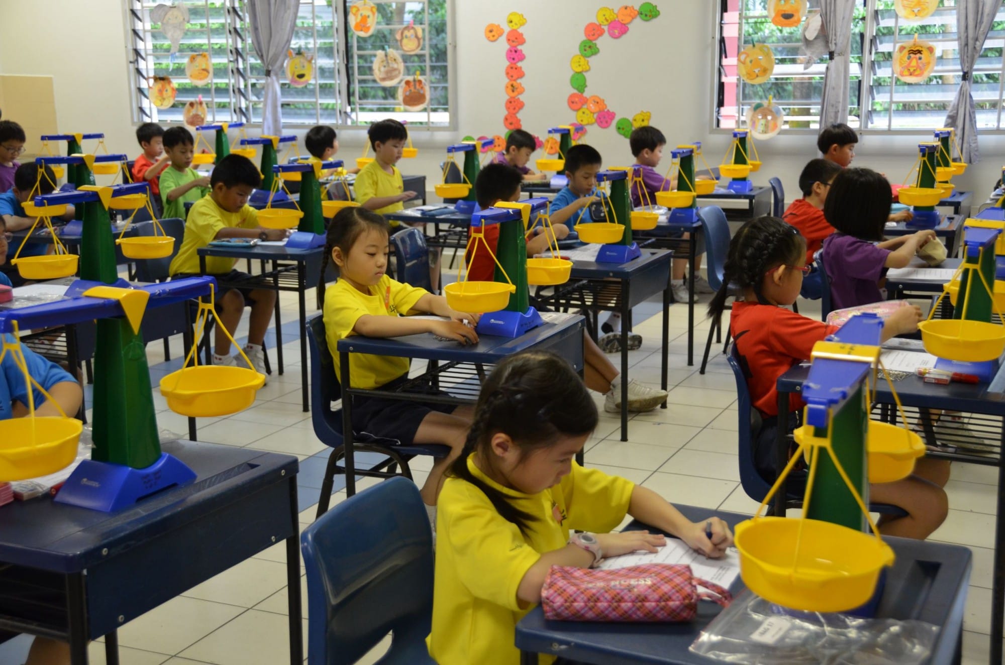 Young children in a decorated classroom sit at desks, each with a yellow and blue balance scale toy.